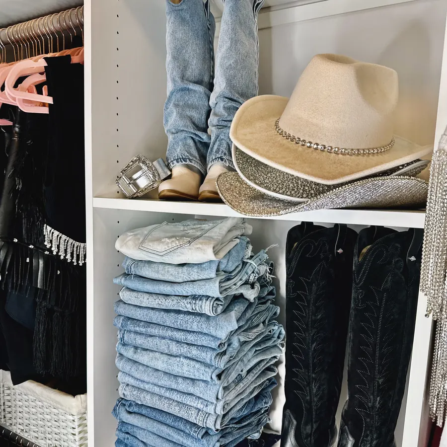 Folded denim stack, cowboy hats, and black western boots on closet shelves